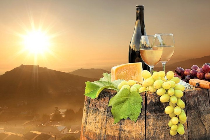 Wine bottle, glasses, grapes, cheese on barrel at sunset with mountain landscape.