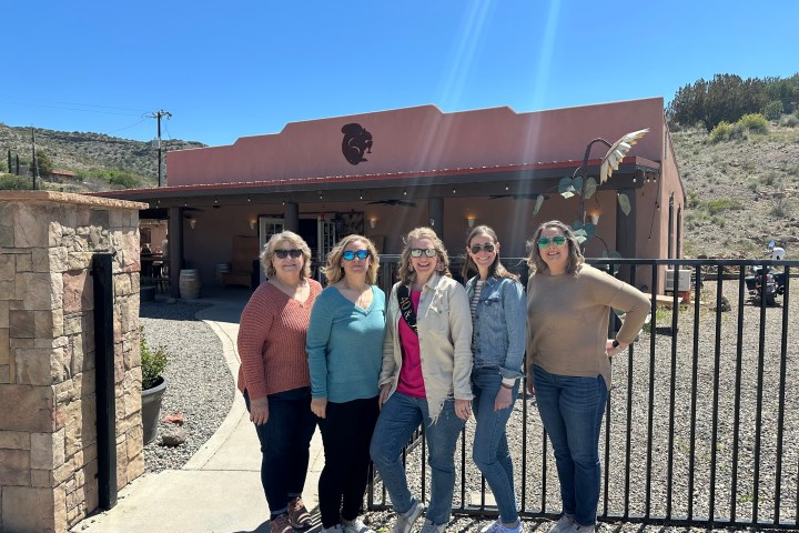 Five women smiling in front of a stucco building on a sunny day.