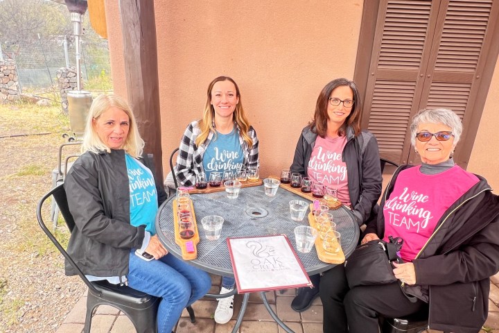 Four women in 'Wine Drinking Team' shirts seated at an outdoor table with drinks.