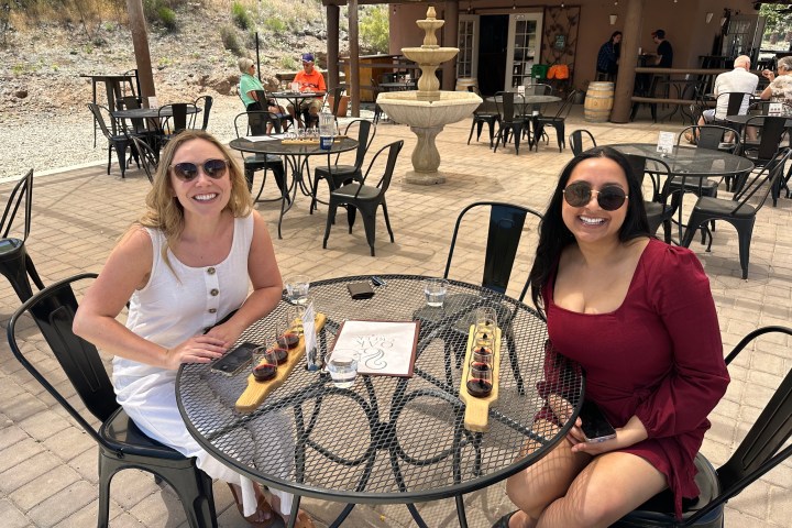 Two women sitting at an outdoor cafe table with wine flights and wearing sunglasses.