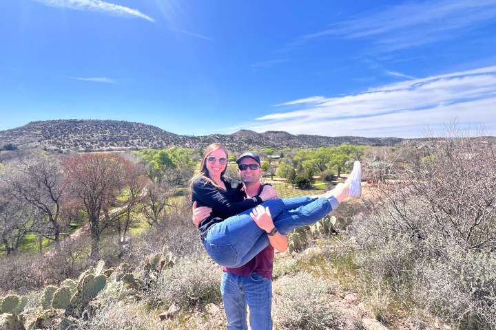 Man in sunglasses holding a woman under a blue sky with desert landscape in the background.