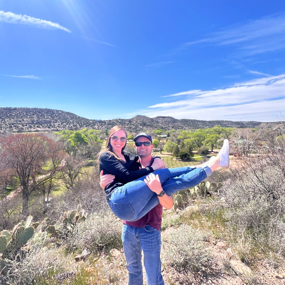 Man in sunglasses holding a woman under a blue sky with desert landscape in the background.