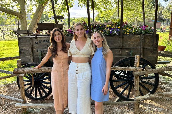 Three women posing in front of a wooden cart with flowers in a park setting.