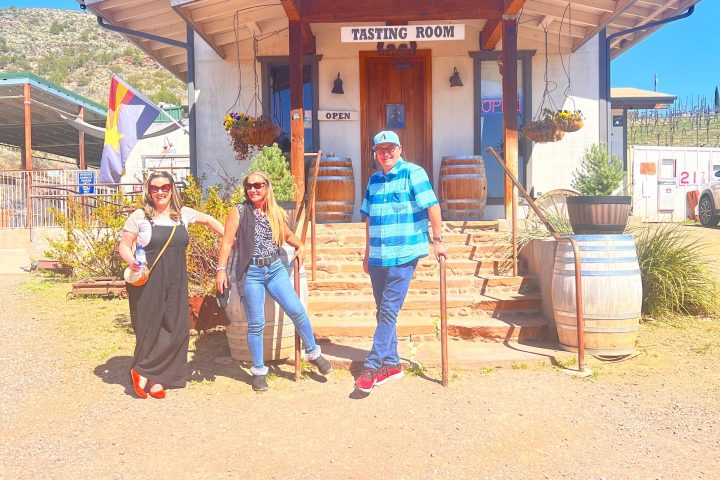 Three people posing outside a rustic tasting room with wooden barrels and a flag, under a clear blue sky.