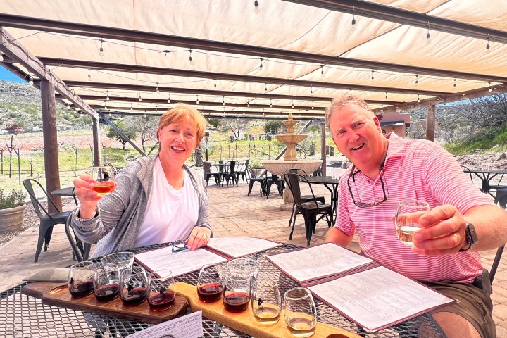 Two people smiling, holding drinks at an outdoor table under a pergola.