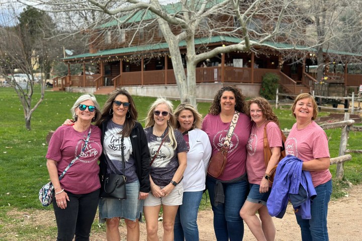 Seven women posing outdoors in front of a large wooden building.
