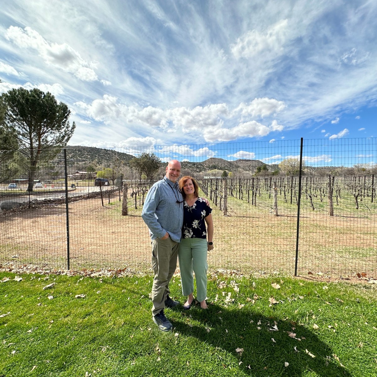 Couple standing on grass near vineyard with cloudy blue sky in background.