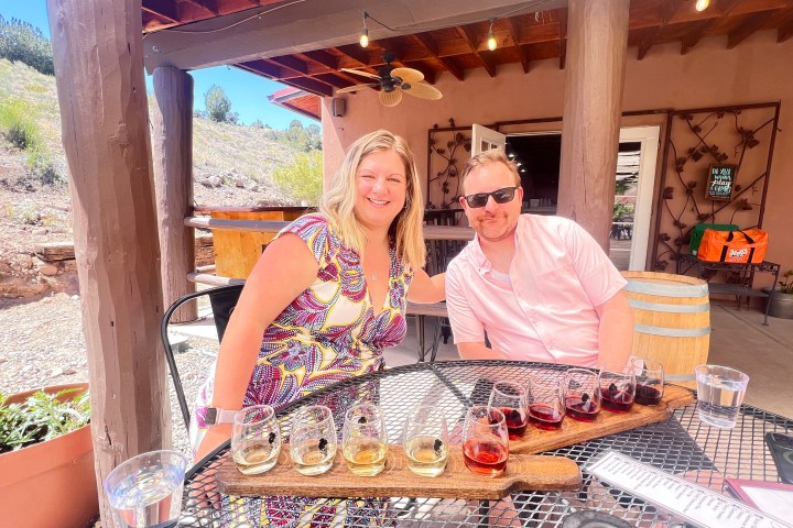 Two people sitting at a winery table outside with wine glasses on the table.