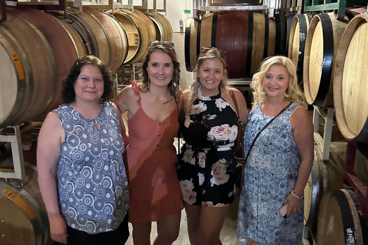 Four women standing in a winery surrounded by stacked wooden barrels.