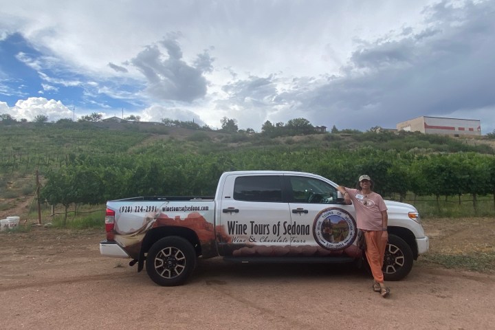 Person leaning on a 'Wine Tours of Sedona' truck in vineyard with cloudy sky.