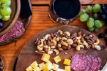 Cheese, nuts, and grapes on a wooden platter with wine glasses on a table.