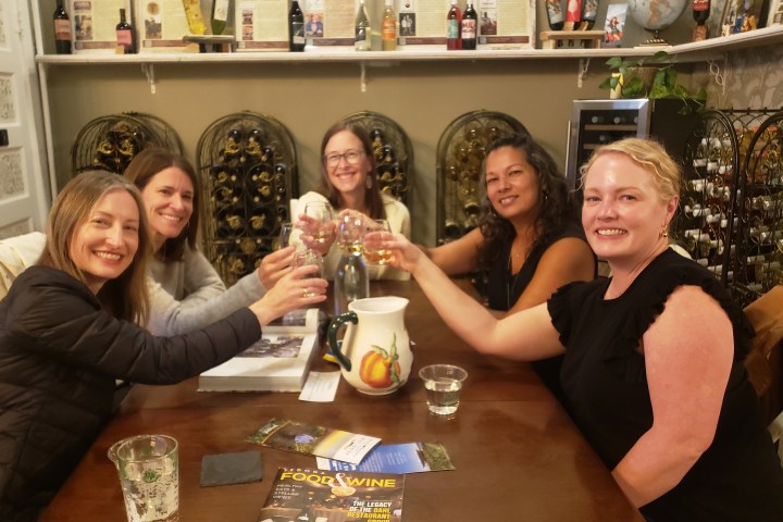 Five women seated around a table, raising glasses in a toast, with wine racks in the background.