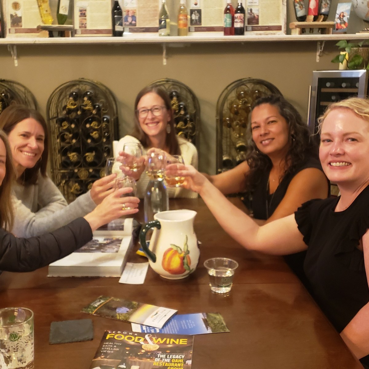 Five women seated around a table, raising glasses in a toast, with wine racks in the background.
