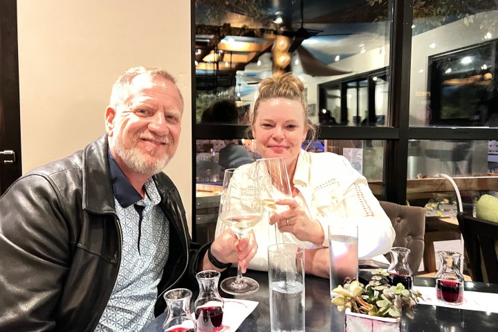 Two people at a restaurant table with wine and drinks, smiling at the camera.