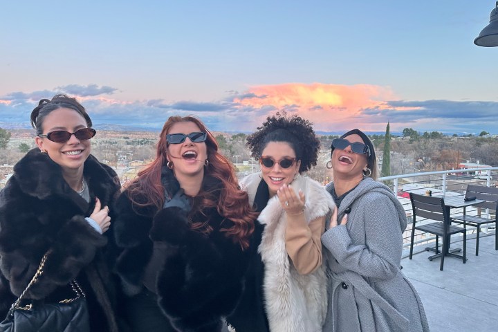 Four women in sunglasses laughing on a balcony with a scenic sunset view.