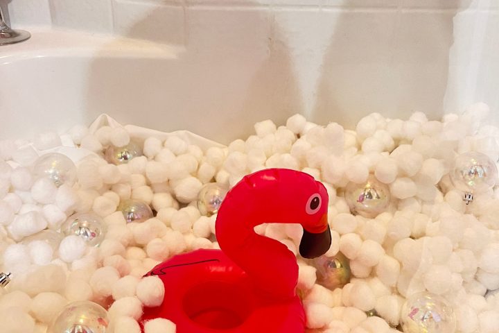 Red inflatable flamingo in a bathtub filled with white cotton balls and clear ornaments.