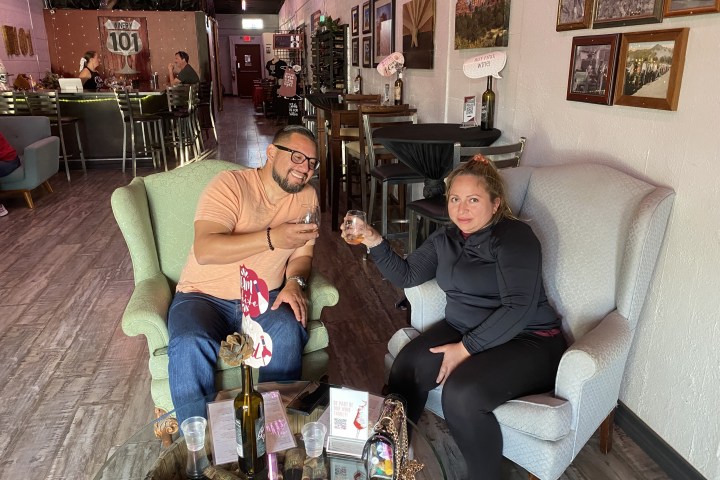 Two people seated in armchairs, holding drinks, in a cozy bar setting with photos on the wall.