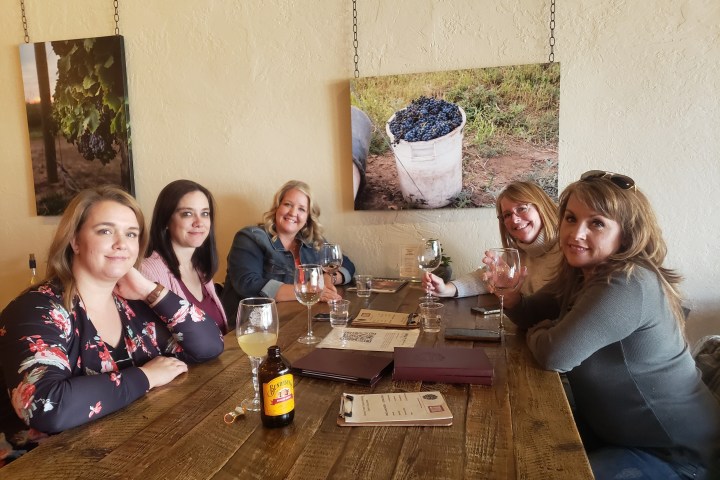 Five women sitting at a wooden table with wine glasses, smiling and looking at the camera.