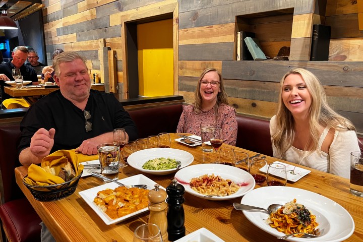 Three people smiling at a restaurant table with various pasta dishes.