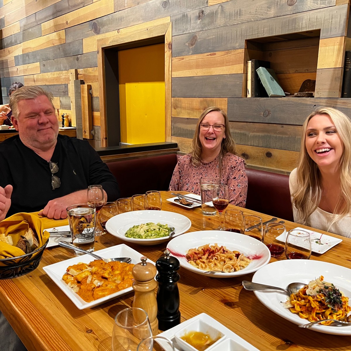 Three people smiling at a restaurant table with various pasta dishes.