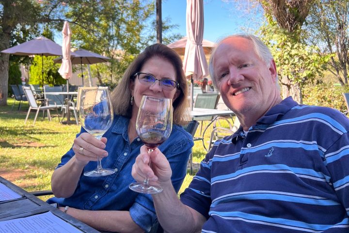 Two people seated outside, smiling and holding wine glasses under a clear blue sky.