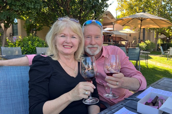 Smiling couple outdoors holding wine glasses, sitting at a table.
