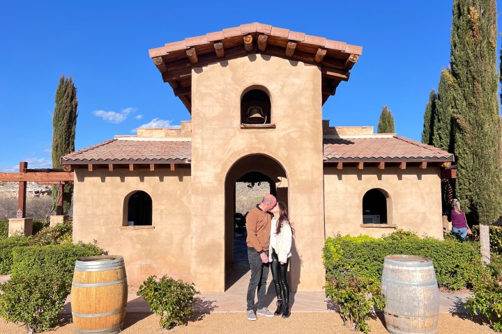 Couple standing in front of a rustic building with a bell tower and cypress trees nearby.