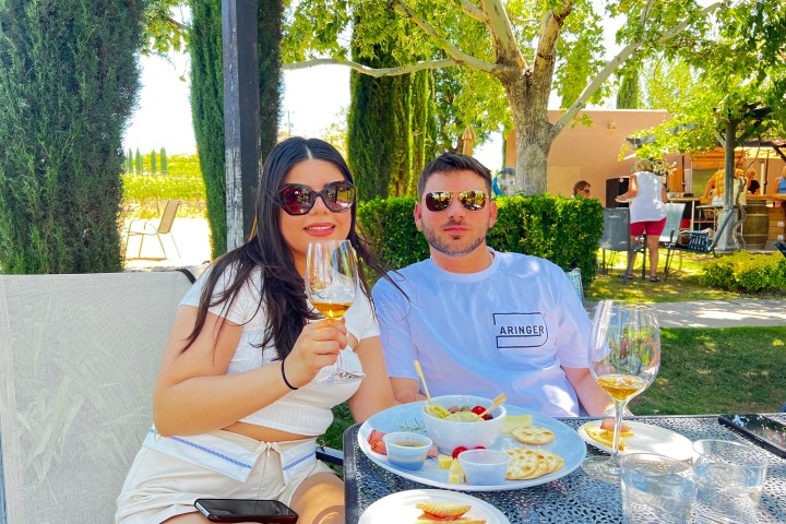Couple sitting outdoors at a table with wine and snacks, surrounded by greenery.