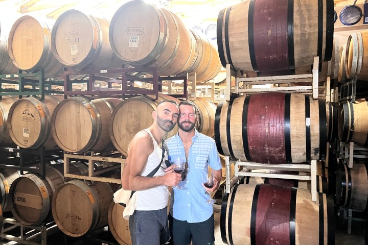 Two people holding wine glasses standing in front of stacked wine barrels in a cellar.