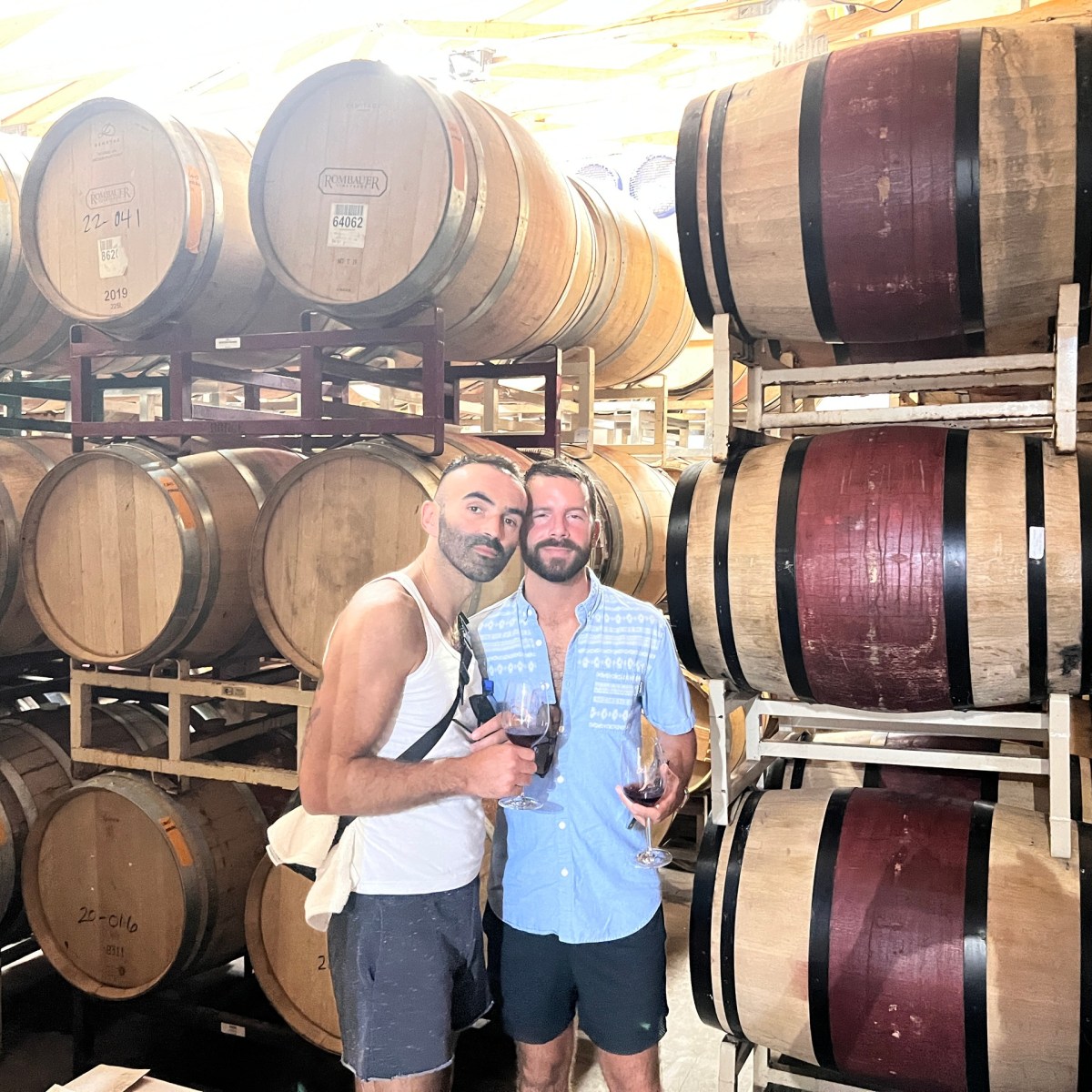 Two people holding wine glasses standing in front of stacked wine barrels in a cellar.