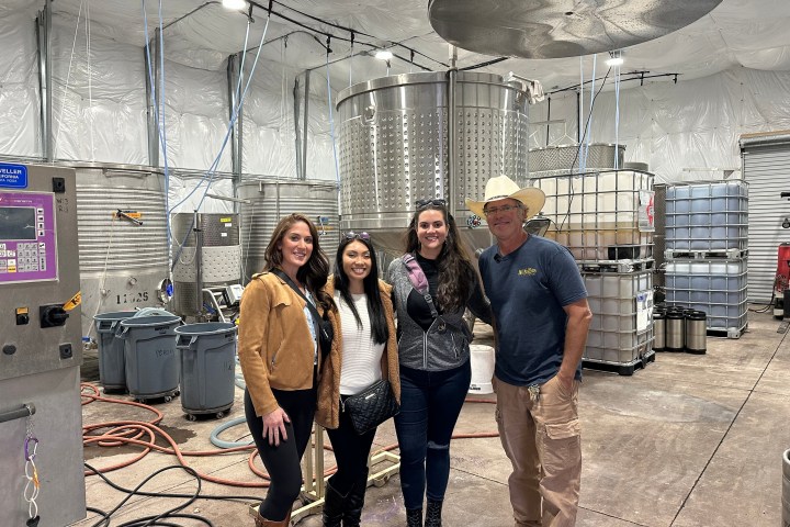 Four people smiling in a winery with large metal tanks and hoses in the background.