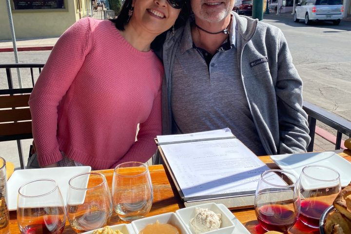 A smiling couple seated at an outdoor table with several wine glasses and appetizers on a sunny day.