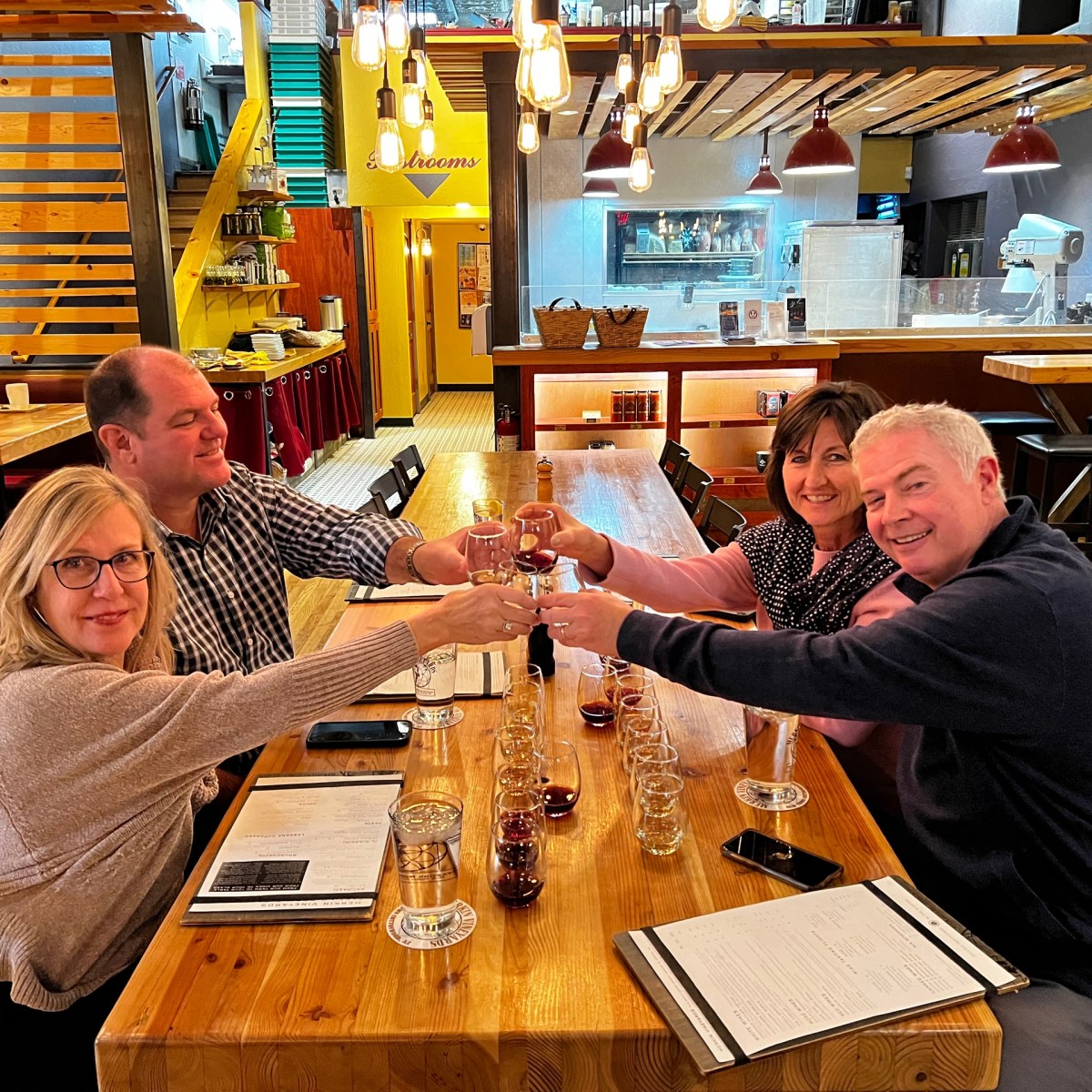 Four people toast with wine glasses at a wooden table in a cozy restaurant.