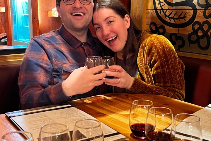 Couple smiling and toasting wine glasses at a wooden table with four glasses of red wine.