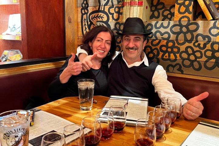 Couple smiling, giving thumbs up at a table with multiple glasses of red wine in a cozy restaurant.