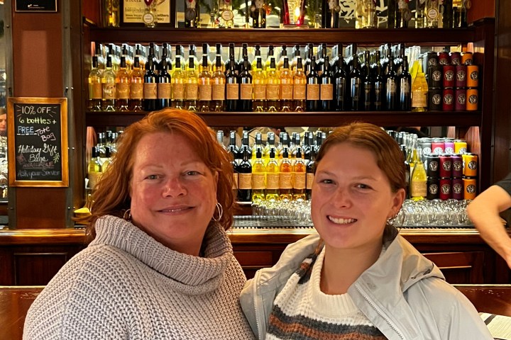 Two people smiling in front of a bar with shelves full of wine bottles.