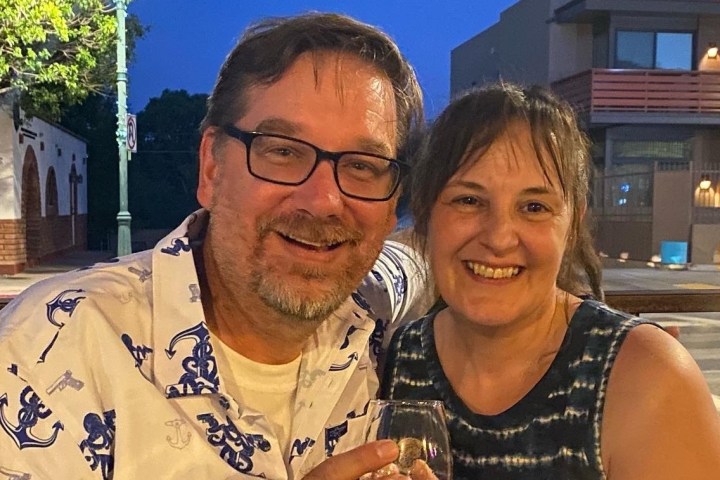 Smiling couple sitting outdoors, holding a drink, with buildings and a streetlamp in the background.