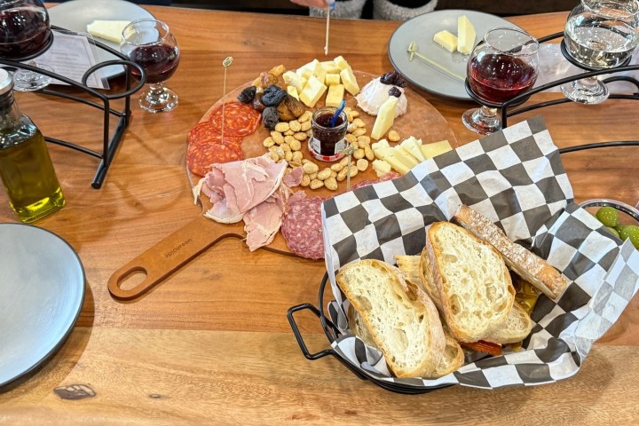 Charcuterie board with meats, cheese, nuts, and bread on a wooden table, surrounded by wine glasses.