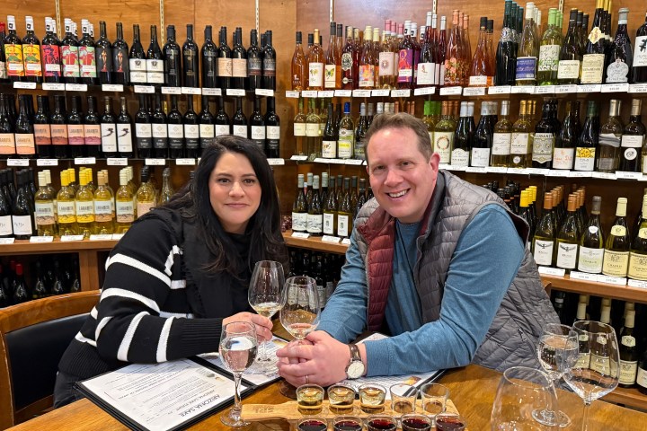 Two people at a wine shop table holding glasses, surrounded by wine bottles on shelves.