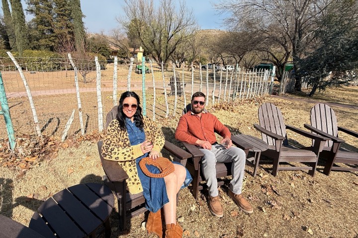Two people sitting on Adirondack chairs in a sunny outdoor setting with trees and a fence.