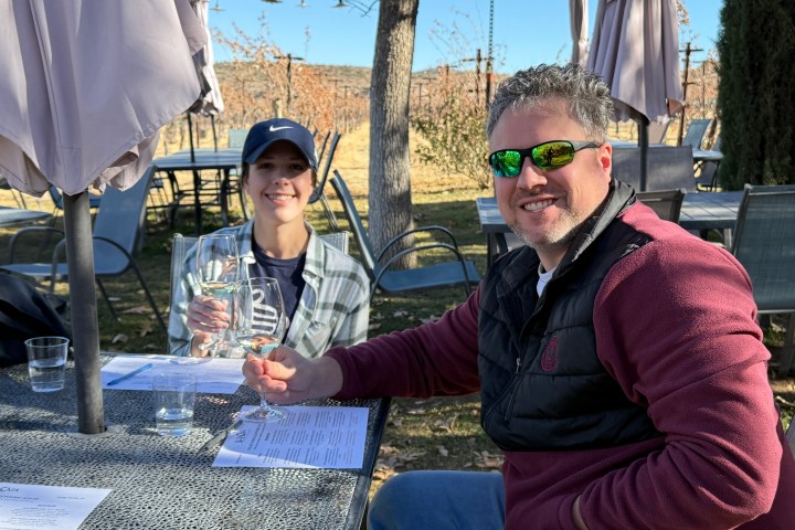 Two people sitting outdoors at a table with drinks, wearing casual attire and smiling.