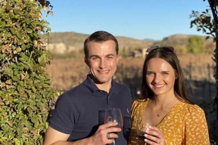 Man and woman holding wine glasses, smiling, standing outdoors with vineyard background and greenery.