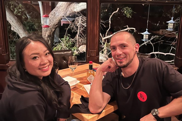 Two people sitting at a restaurant table with tree visible outside window.
