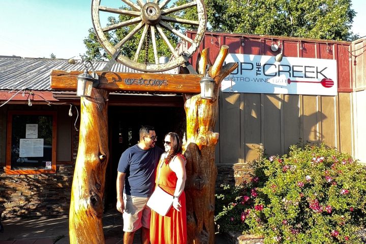 Couple stands under wheel arch at 'Up The Creek' sign; sunny day with greenery.