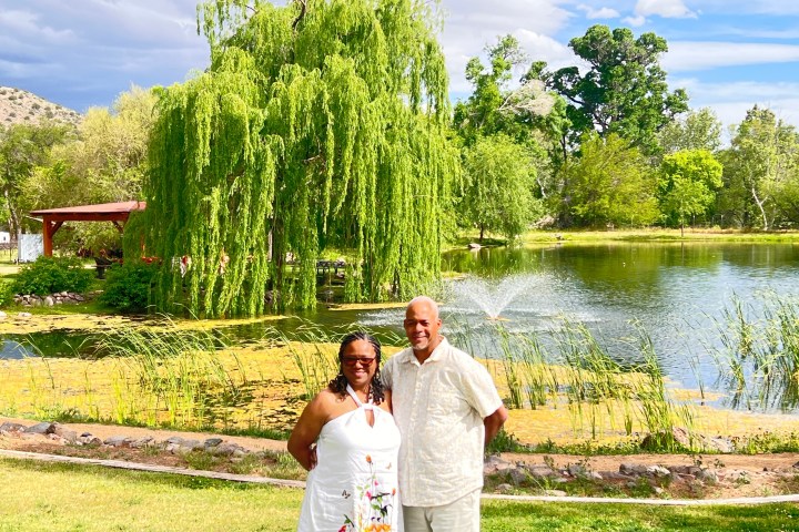 A couple stands in front of a pond with a fountain and lush greenery.