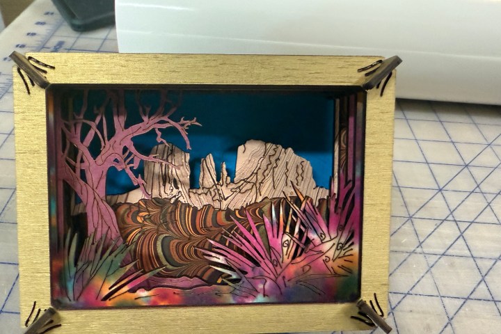 Colorful layered paper art depicting a tree, mountains, and plants in a wooden frame.