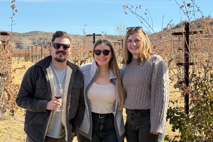 Three people standing in a vineyard, smiling at the camera; one holds a glass of red wine.