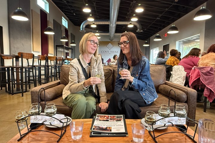 Two women sitting on a couch in a winery, holding wine glasses, with people in the background.
