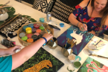 Two people painting colorful tropical scenes, surrounded by paints and brushes on a table.