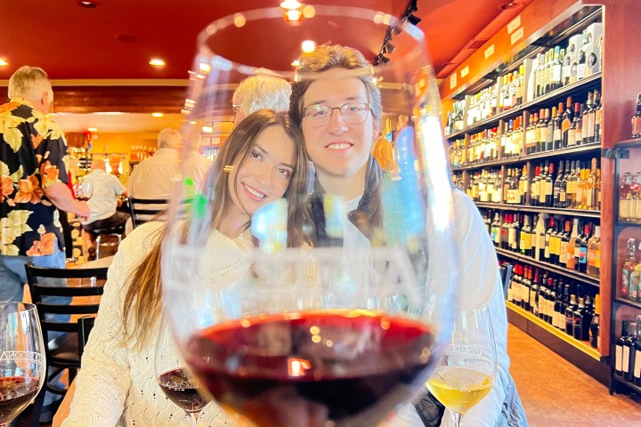 A couple smiles behind a glass of red wine in a wine shop filled with bottles.
